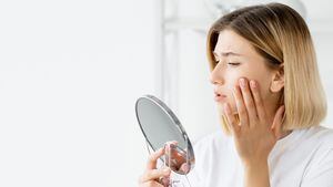 Problema de la piel. Tratamiento contra el acné. Atención médica dermatológica. Perfil de mujer triste frustrada tocando la cara en el espejo sobre fondo interior blanco desenfocado.