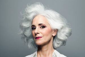 Mature woman with medium length, big, wavy, silvery, grey hair in front of grey background wearing make up red lip stick, portrait.