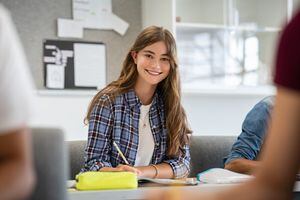 Hermosa estudiante universitaria sonriendo mientras estudiaba para los exámenes en el aula. Linda mujer sentada en un aula llena de estudiantes durante la clase. Retrato de una joven feliz escribiendo notas y mirando a la cámara.