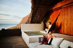 Wide shot of gay couple relaxing on deck of luxury tropical villa drinking coffee and watching sunrise