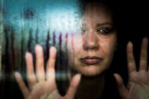 Cierre la imagen en color desaturado que representa a una mujer triste y deprimida de unos 30 años, y de etnia caucásica, mirando una ventana empapada de lluvia con las manos presionadas contra el cristal. La imagen se tomó durante la pandemia de Covid-19 durante el encierro, y la imagen ilustra un concepto de salud mental, así como un concepto de abuso doméstico. Habitación para espacio de copia.
