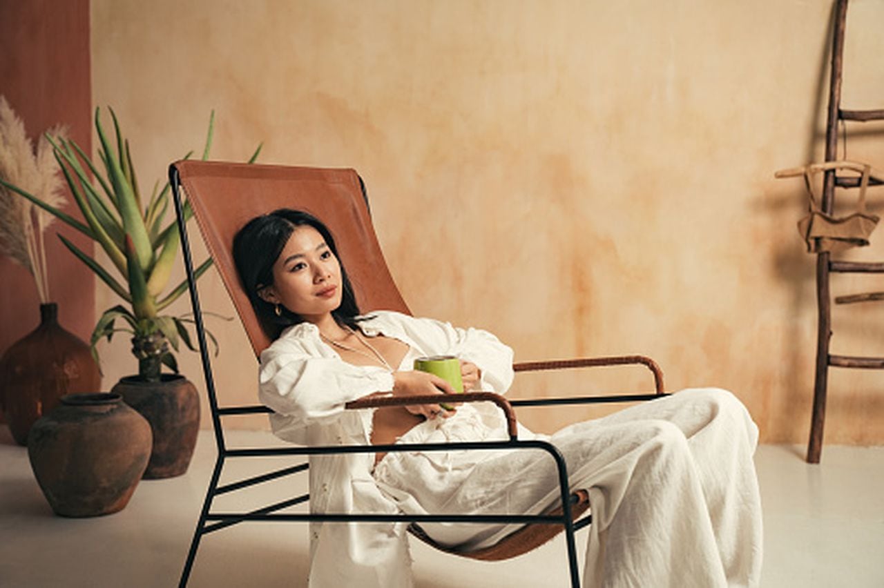 Gorgeous chinese woman in white bra and pajama lying on lounger with rustic interior on background, holding cup of hot relaxing drink in hands, looking aside, spending morning in spa resort
