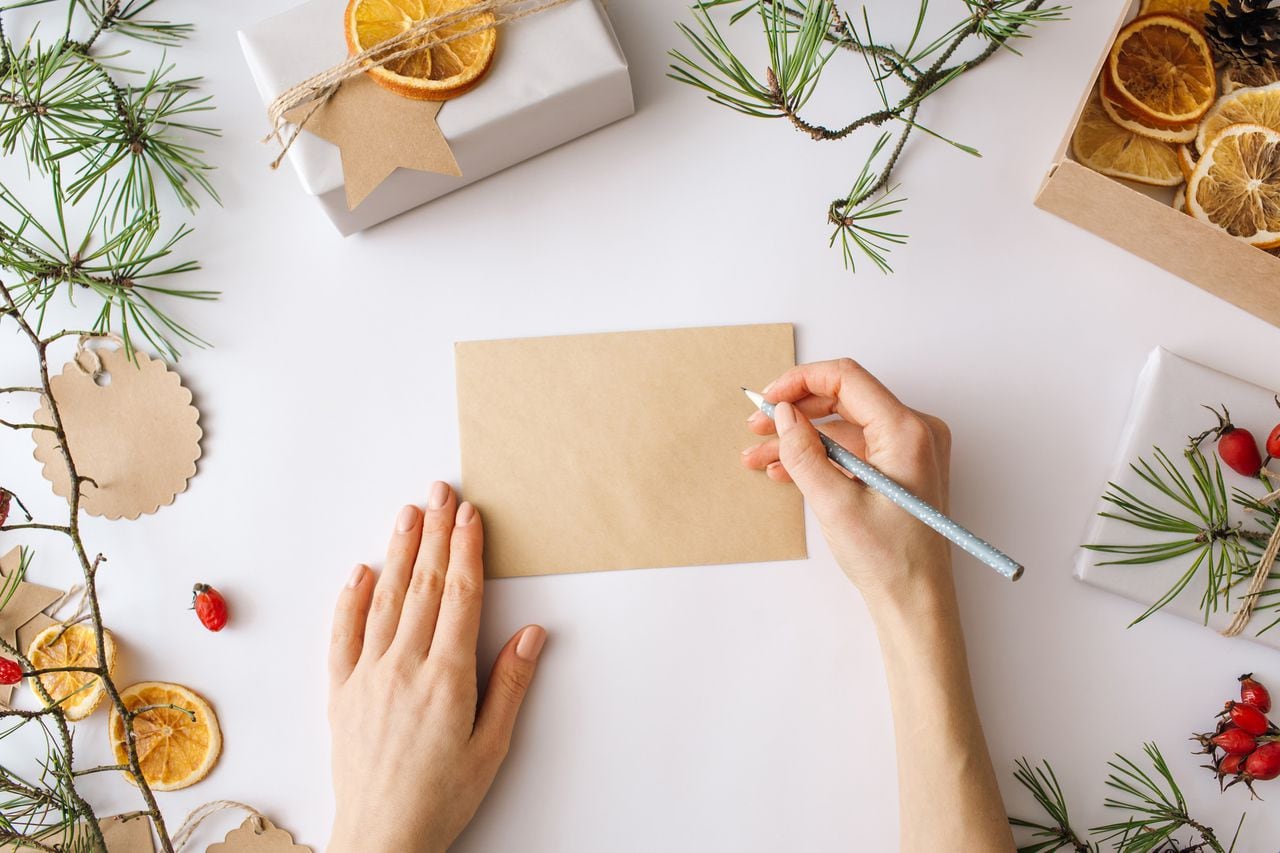 Christmas blank greeting card mock-up. Woman writing in a Blank Letter Festive winter composition. Craft envelope, gift boxes, nature decorations on white background. Flat lay, top view, copy space.
