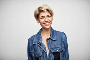 Portrait of smiling young woman in denim jacket. Fashionable Latin American female with blond short hair. She is against white background.