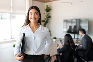 Portrait of hispanic businesswoman with a file standing in meeting room with colleagues disucssing in background