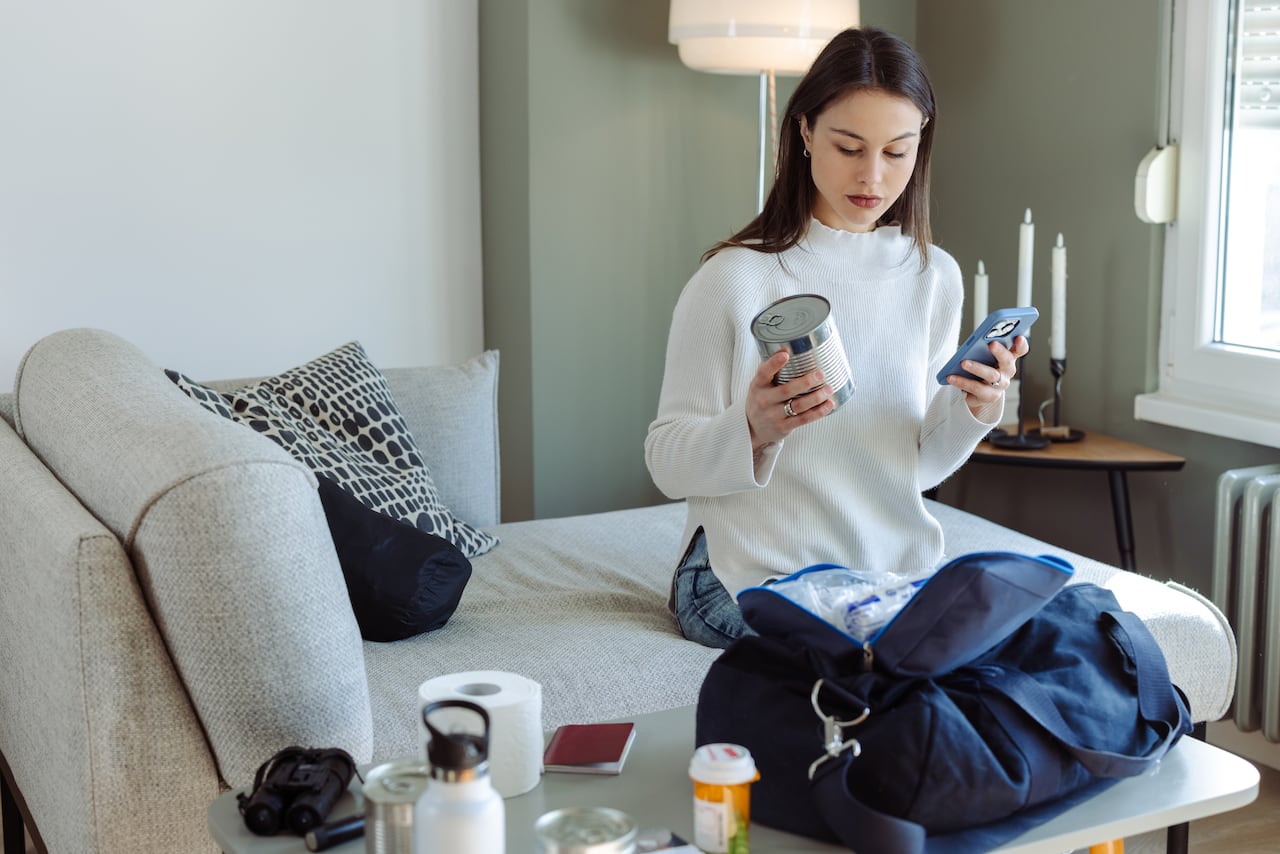 Photo of a young woman holding smartphone and canned food while packing essentials in bag