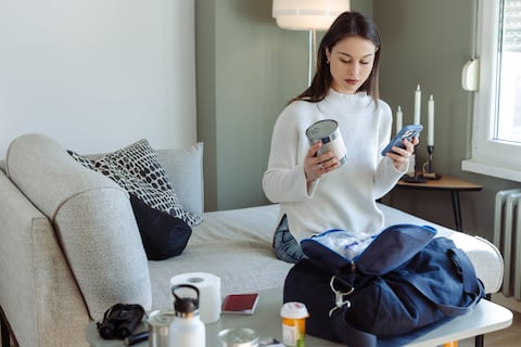 Photo of a young woman holding smartphone and canned food while packing essentials in bag
