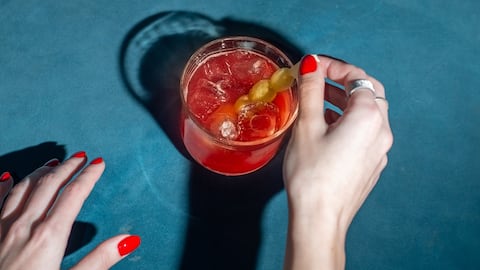 Female hand with red nails and red cocktail on turquoise velvet background. Nightlife and mixology concept
