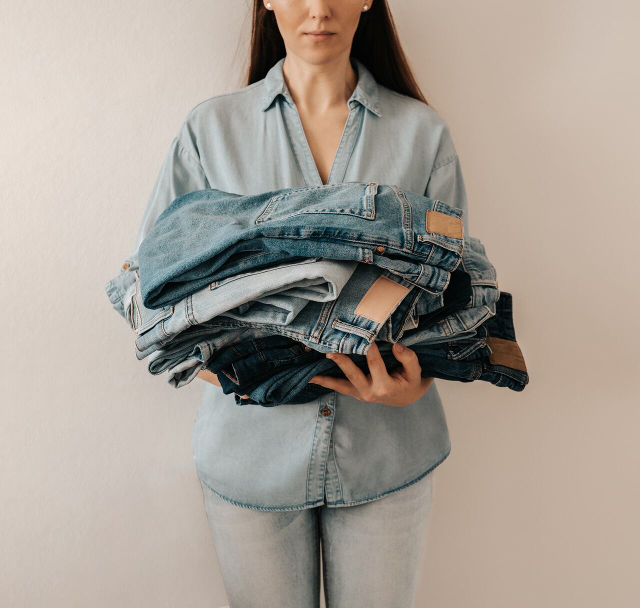 Unrecognizable woman holds stack of lot jeans pants on white background. Caucasian woman with long brown hair weared in blue shirt and jeans holds heap of denim pants with differents shades of blue