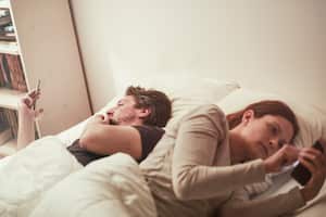 Man and woman lying in bed, each on his or hers side, on the smart phone. Emotional emptiness.