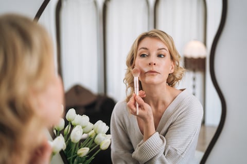 Beautiful adult blonde woman applying blush makeup at a mirror in her bedroom. Beauty and daily routine concept