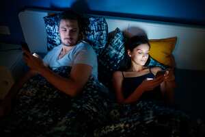 Young couple lying in bed in the evening with mobile phones, man looking at woman's phone with suspicion