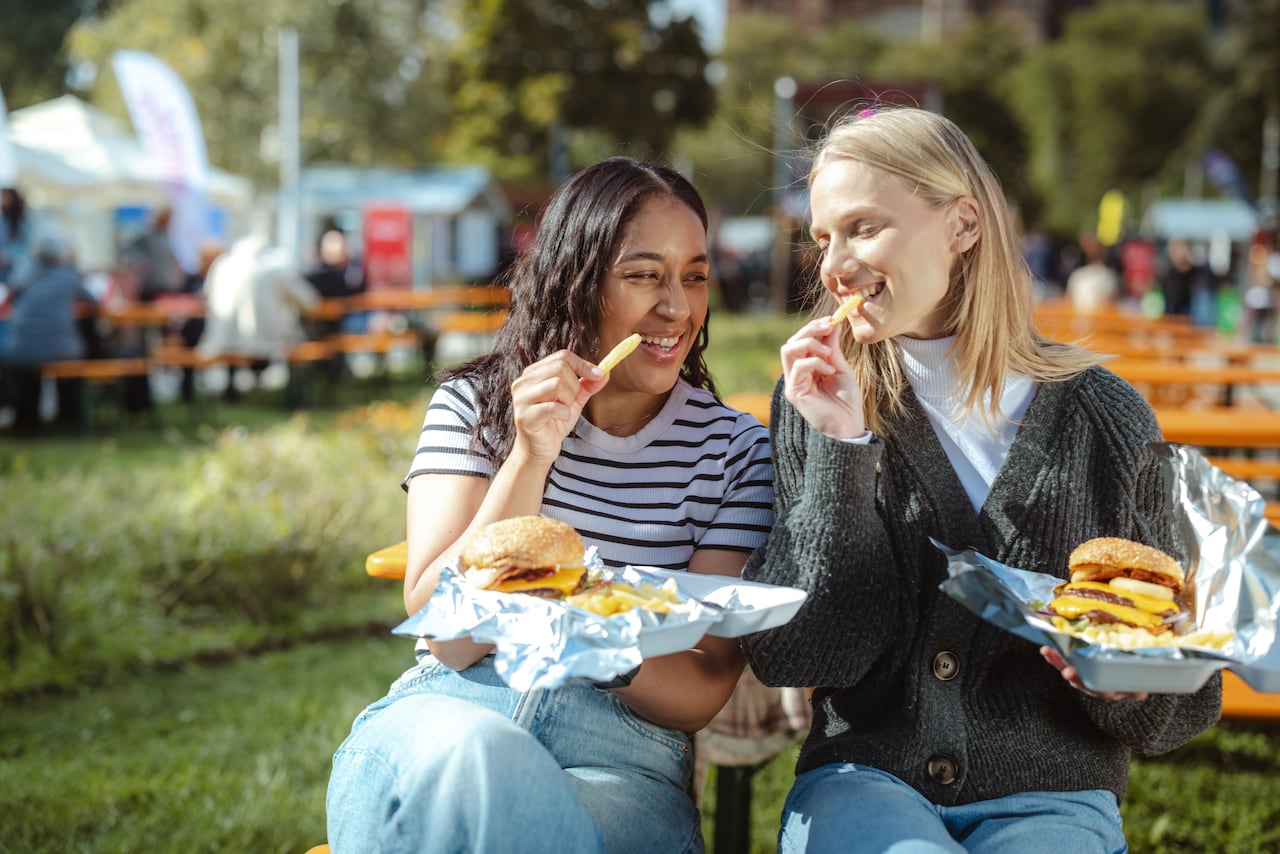 Two friends enjoying burgers at an outdoor festival in a sunny park setting