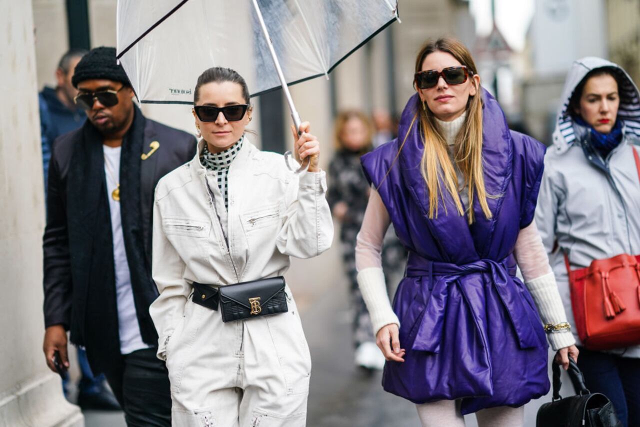 PARIS, FRANCE - FEBRUARY 28: Julia Comil (L) wears sunglasses, a grey and black checkered turtleneck, a white jumpsuit, a black Burberry belt bag with a coin purse, and holds an umbrella  ;  Natalia Ostrofsky (R) wears sunglasses, one earring, bracelets, a white ribbed turtleneck with transparent sleeves, a purple puffer vest, white tights, outside Ralph & Russo, during Paris Fashion Week - Womenswear Fall/Winter 2020/2021, on February 28, 2020 in Paris, France. (Photo by Edward Berthelot/Getty Images)