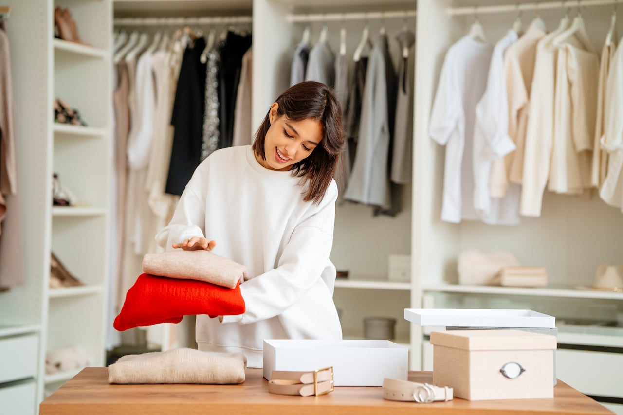 Brunette woman packing and stacking clothes items into the boxes in a big showroom or wardrobe room with a lot of clothing and shoes racks