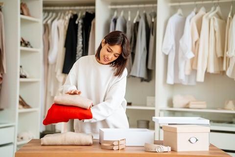 Brunette woman packing and stacking clothes items into the boxes in a big showroom or wardrobe room with a lot of clothing and shoes racks