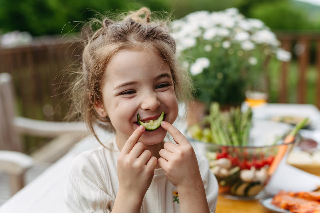 Young girl eating slice of cucumber, having fun, waiting for grilled food. Family barbecue in backyard, spending quality time outdoors together.