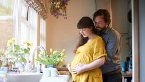 A pregnant couple embrace with there hands gently on stomach in the kitchen of their home with fresh spring flowers in vase.