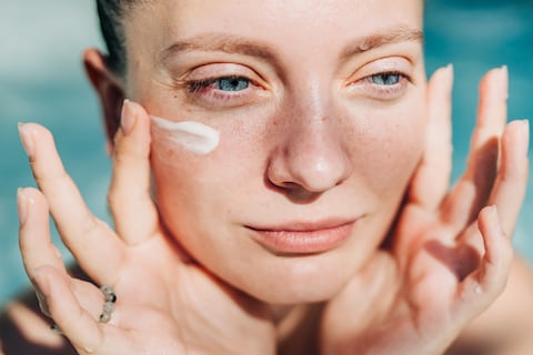 A close-up portrait of a woman with her eyes closed and a serene expression, as she applies a small dab of sunscreen or face cream to her cheek.