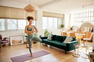 Full length of fit woman practicing tree pose at home. Young woman standing on one leg on exercise mat. She is doing yoga in living room.